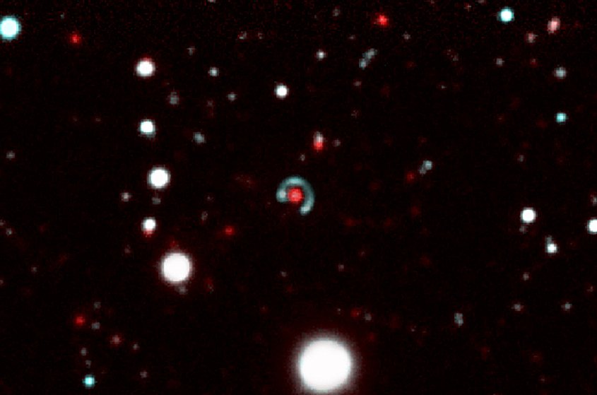 An image of the "Canarias Einstein ring". In the centre of the picture, we can see how the source galaxy (the greenish-blue circle), which is further away "surrounds" the lens galaxy (red dot) almost completely. This phenomenon is produced because the strong gravitational field of the lens galaxy distorts the space-time in its neighborhood, bending the paths of the light rays which come from the source galaxy. As they are almost perfectly aligned, the resulting image of the source galaxy is almost circular. Credit: Image made up from several images taken with the DECam camera on the Blanco 4m telescope at the Cerro Tololo Observatory in Chile